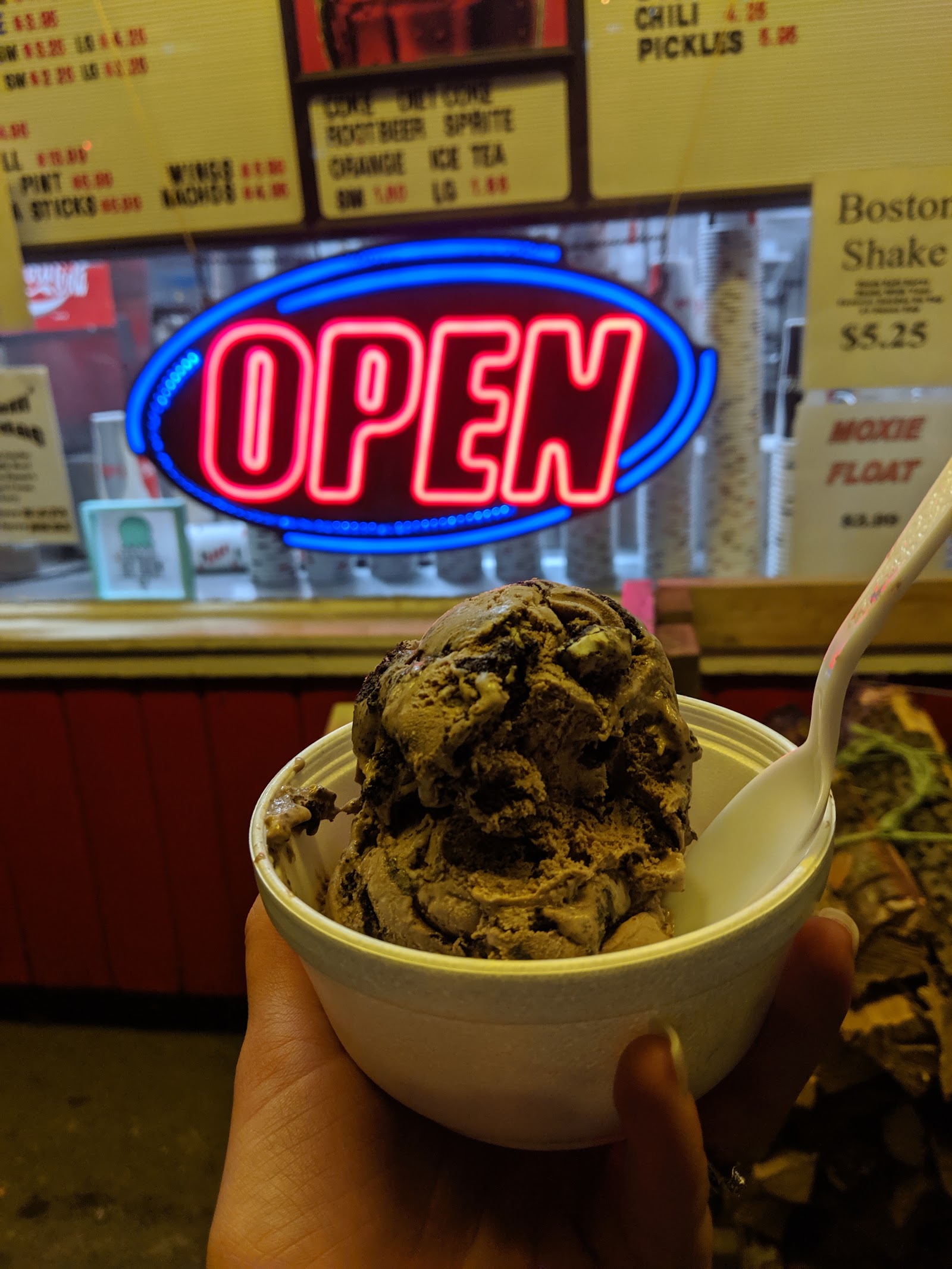 A bowl of chocolate hard‑serve ice cream held up in front of the glowing neon OPEN sign at night.