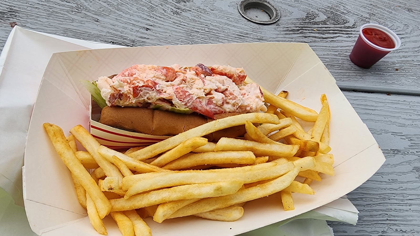 A lobster roll packed with lobster salad beside a heap of french fries in a paper boat.