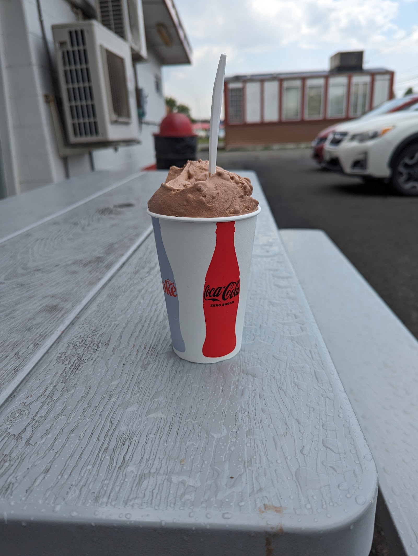 A chocolate soft‑serve in a Coca‑Cola paper cup on a picnic table, with the stand behind.