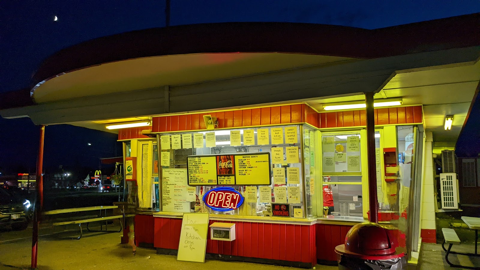 The red‑and‑cream Sawyer’s stand lit up at dusk, its printed menu board glowing and the neon OPEN sign on.