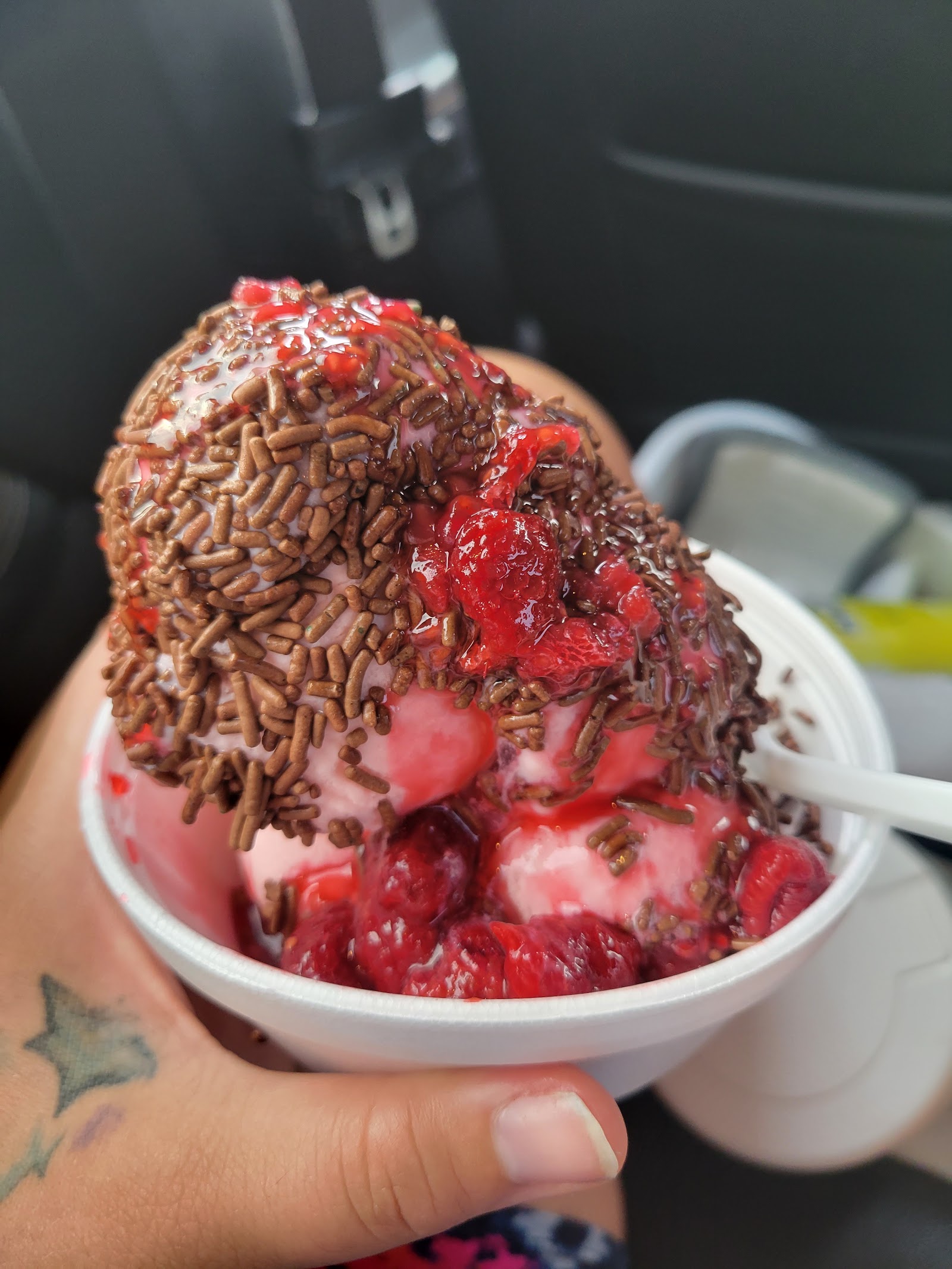 A strawberry sundae topped with chocolate jimmies and strawberry sauce in a paper cup.